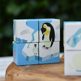 A set of Bajo cube puzzle wooden toy blocks featuring an arctic penguin design, displayed on a rustic wooden slice with a blurred green outdoor background.