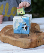 A child's hand is placing a colorful animal block onto a stack of wooden educational blocks featuring polar animals like a penguin and polar bear, set on a rustic wooden serving board.
