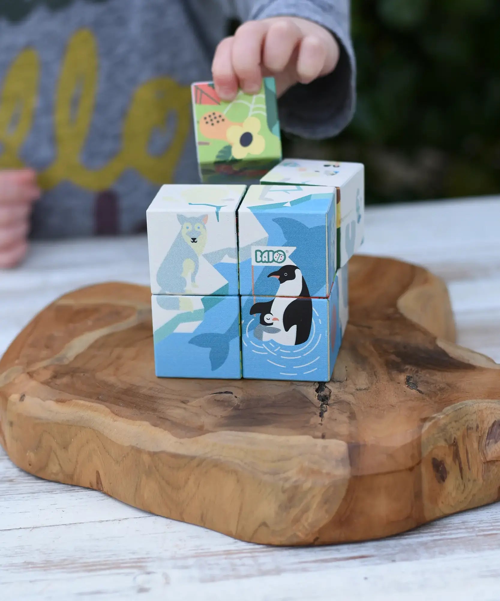 A child's hand is placing a colorful animal block onto a stack of wooden educational blocks featuring polar animals like a penguin and polar bear, set on a rustic wooden serving board.