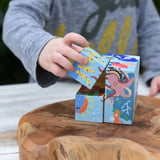 A child's hand taking a cube from the Bajo 2-in-1 reef desert wooden cube block puzzle. Colorful wooden toy cube with sea creature illustrations on one side and desert scenes on the other.