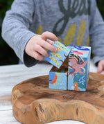 A child's hand taking a cube from the Bajo 2-in-1 reef desert wooden cube block puzzle. Colorful wooden toy cube with sea creature illustrations on one side and desert scenes on the other.