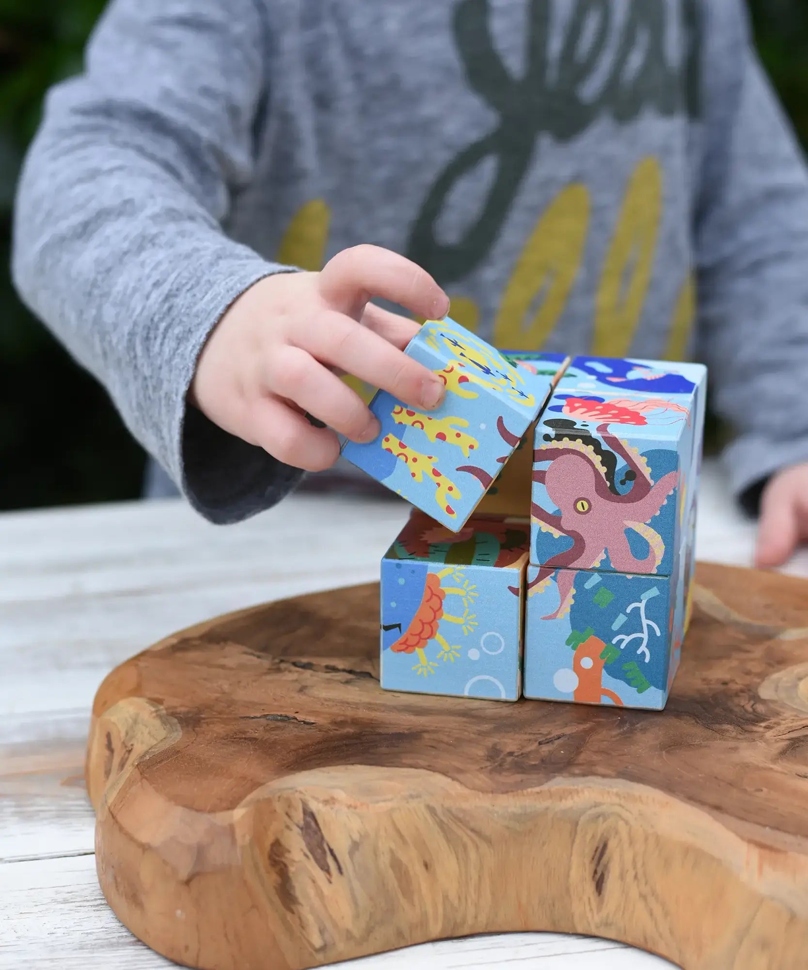 A child's hand taking a cube from the Bajo 2-in-1 reef desert wooden cube block puzzle. Colorful wooden toy cube with sea creature illustrations on one side and desert scenes on the other.