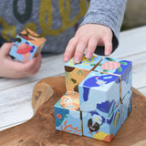 Child's hands playing with colorful wooden puzzle blocks from the Bajo 2-in-1 cube reef desert puzzle on a rustic wooden board. The blocks feature illustrated scenes of sea life and nature.