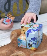 Child's hands playing with colorful wooden puzzle blocks from the Bajo 2-in-1 cube reef desert puzzle on a rustic wooden board. The blocks feature illustrated scenes of sea life and nature.