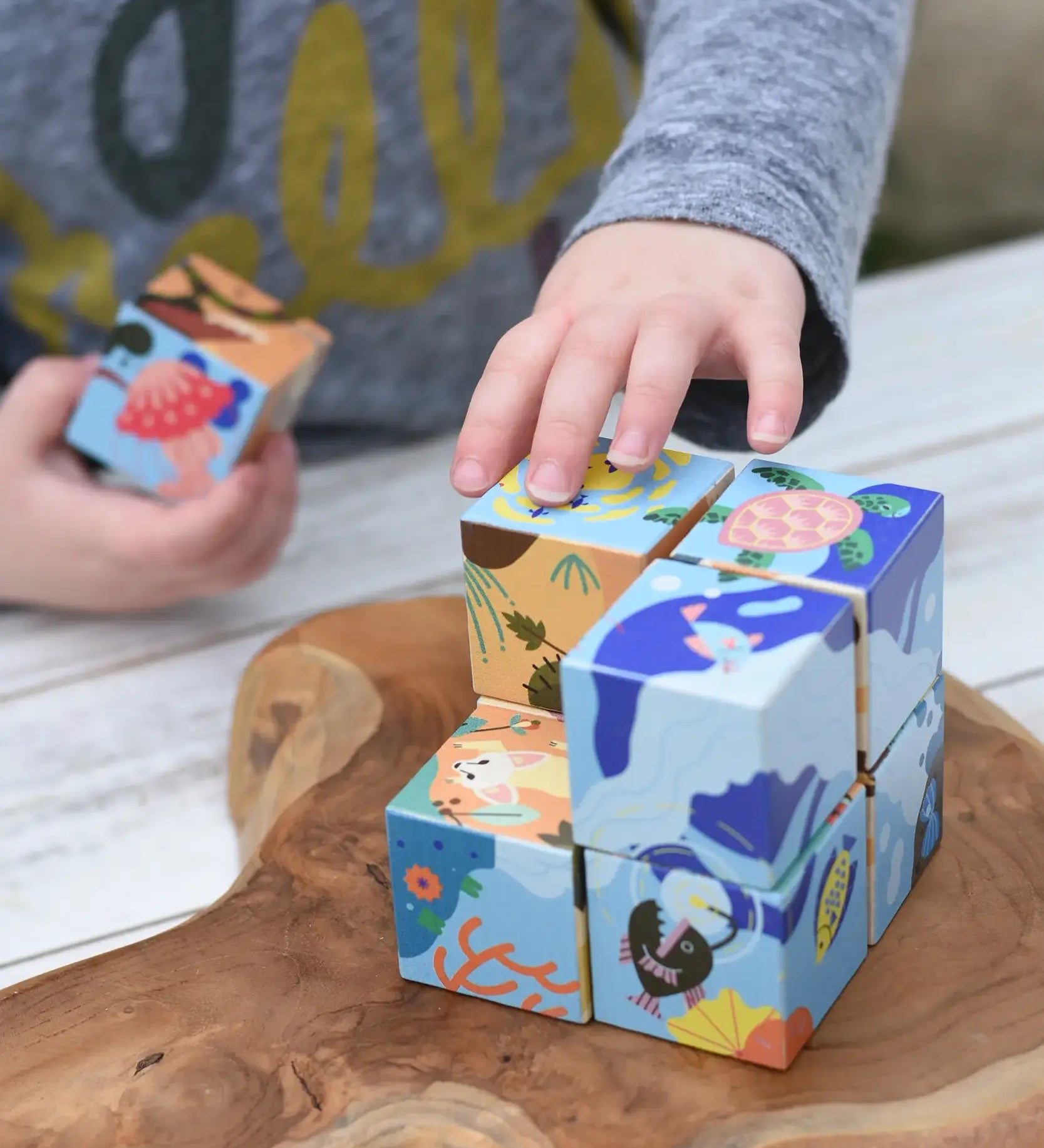 Child's hands playing with colorful wooden puzzle blocks from the Bajo 2-in-1 cube reef desert puzzle on a rustic wooden board. The blocks feature illustrated scenes of sea life and nature.