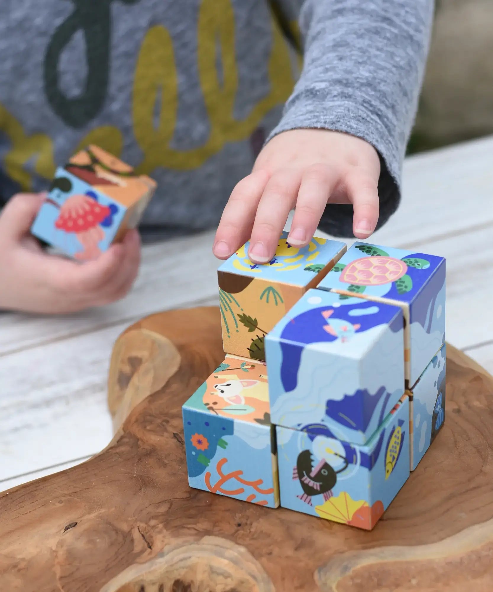Child's hands playing with colorful wooden puzzle blocks from the Bajo 2-in-1 cube reef desert puzzle on a rustic wooden board. The blocks feature illustrated scenes of sea life and nature.