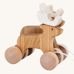 A side on view of the Bajo Pull Along Jumping Reindeer, showing the natural wood and white antlers of the reindeer. The image also shows how the two reindeer's are attached together by two wooden wheels. The image is on a cream background