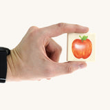 An adult hand holding a square tile from the Bajo classic memory game available at Babipur showing an apple illustration and the scale of the tile