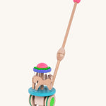 The Bajo Push Along Carousel has a long wooden handle with a pink wooden ball on top. It also has wooden horses which spin around when the toy is pushed along the ground. The Carousel is blue, purple, green, pink and natural wood in colour. The image is on a cream background