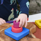 Child's hand placing the cylinder pieces from the Bajo Puzzle Stacker Tower on the large blue square base piece.