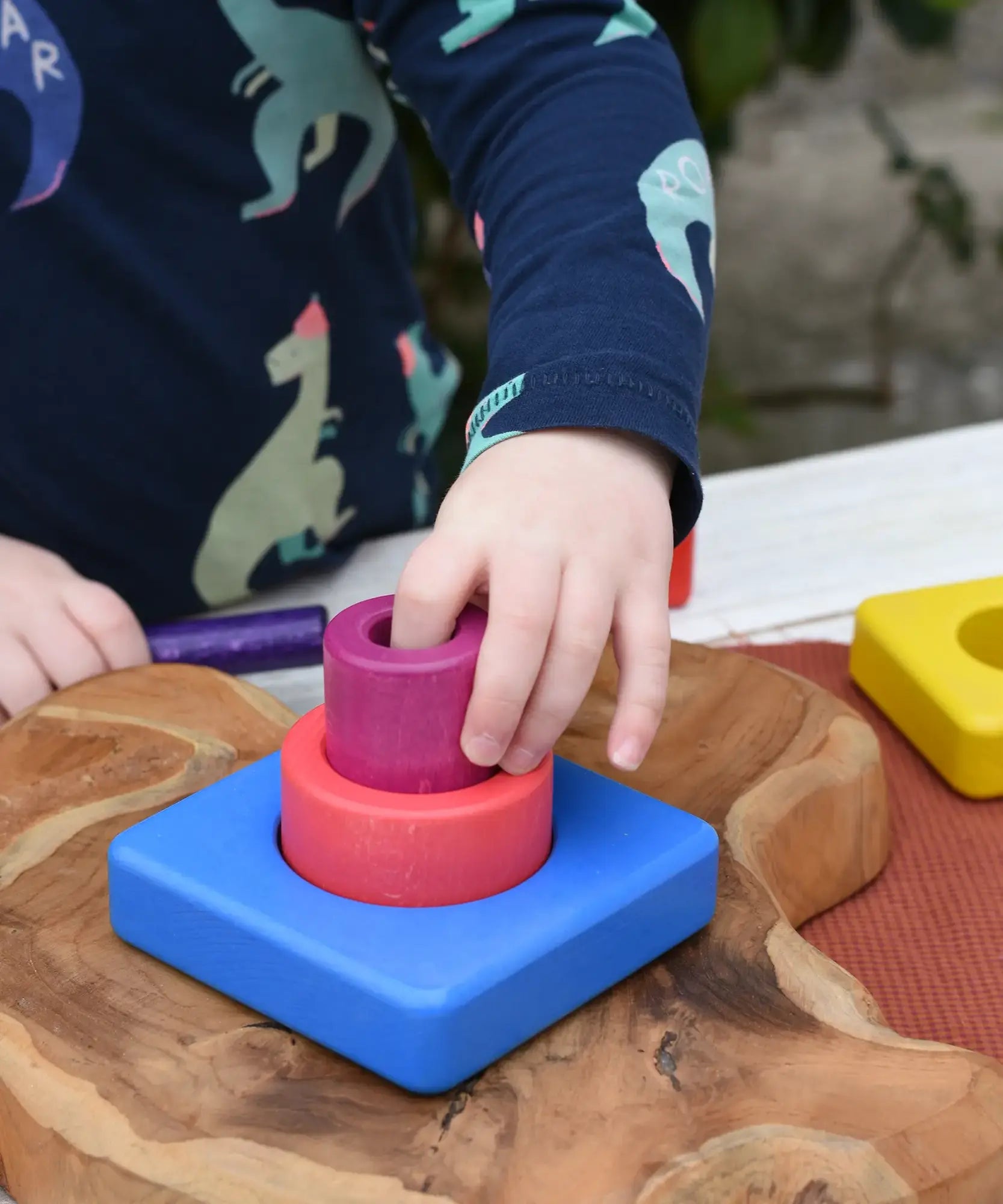Child's hand placing the cylinder pieces from the Bajo Puzzle Stacker Tower on the large blue square base piece.