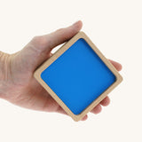 An adult hand holding the biggest wooden square block from the Bajo squaremide sensory blocks available at Babipur. This block has a blue base
