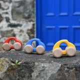 A group of Bajo handmade wooden beetle style car including blue, yellow and red available at Babipur. These have unpainted wheels and can be pushed or pulled along - photo on a stone wall.