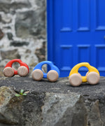 A group of Bajo handmade wooden beetle style car including blue, yellow and red available at Babipur. These have unpainted wheels and can be pushed or pulled along - photo on a stone wall.