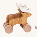 Bajo Wooden Pull Along Jumping Reindeer, is a beautiful children's toy of two natural wooden reindeer with wooden wheels and white antlers, and a wooden rectangle piece to pull along the reindeer which is attached to the reindeer with white string. The image is on a cream background