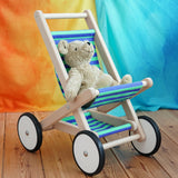 A BAJO Toys wooden stroller buggy with a striped blue and green fabric seat, featuring a beige Senger organic teddy bear sitting inside, set on a wooden floor against a colorful orange, yellow, and blue backdrop.