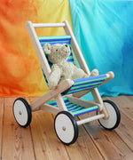 A BAJO Toys wooden stroller buggy with a striped blue and green fabric seat, featuring a beige Senger organic teddy bear sitting inside, set on a wooden floor against a colorful orange, yellow, and blue backdrop.