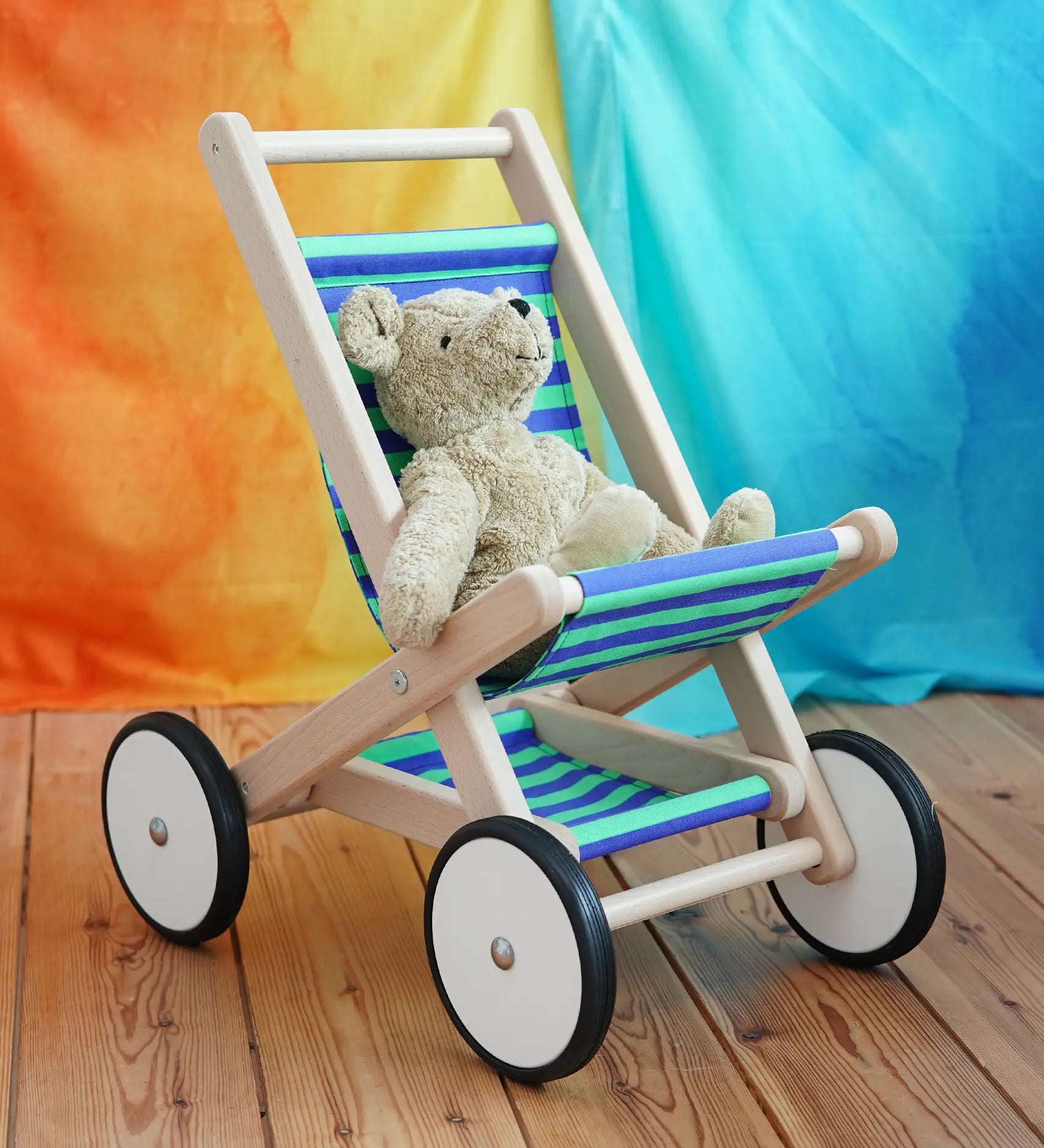 A BAJO Toys wooden stroller buggy with a striped blue and green fabric seat, featuring a beige Senger organic teddy bear sitting inside, set on a wooden floor against a colorful orange, yellow, and blue backdrop.