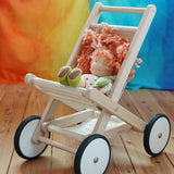 Bajo natural wood toy stroller with four wheels, styled with a kathe Kruse doll that has orange hair and green shoes, set on a wooden floor against a colorful orange, yellow, and blue background.