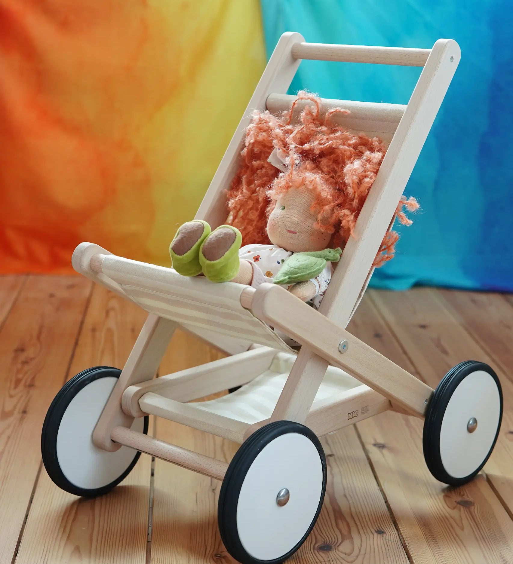 Bajo natural wood toy stroller with four wheels, styled with a kathe Kruse doll that has orange hair and green shoes, set on a wooden floor against a colorful orange, yellow, and blue background.