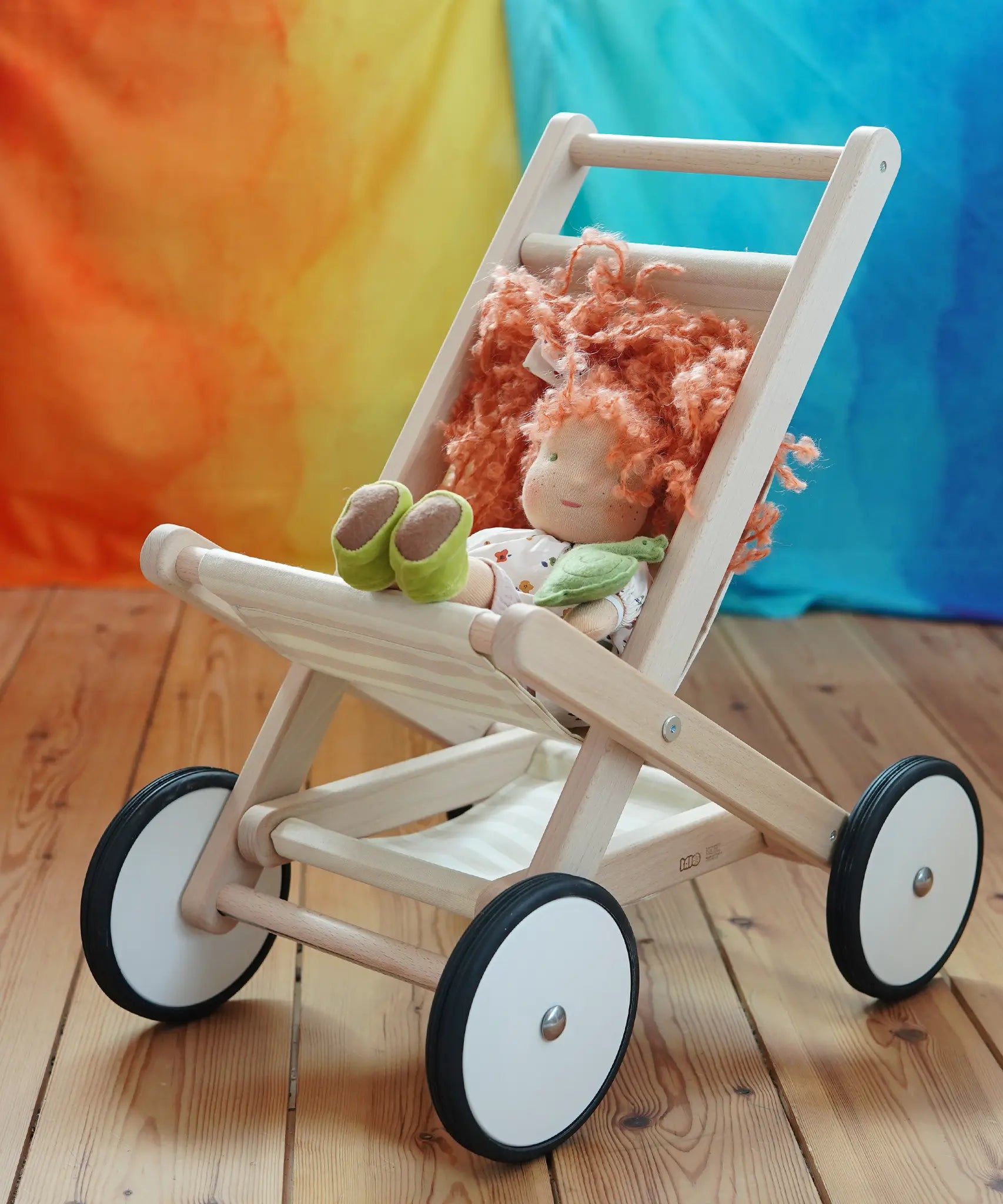 Bajo natural wood toy stroller with four wheels, styled with a kathe Kruse doll that has orange hair and green shoes, set on a wooden floor against a colorful orange, yellow, and blue background.