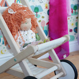 A wooden Bajo Toys stroller with a doll featuring curly orange hair and a white dress, set against a colorful children's room background with patterned wallpaper and a pink blanket.