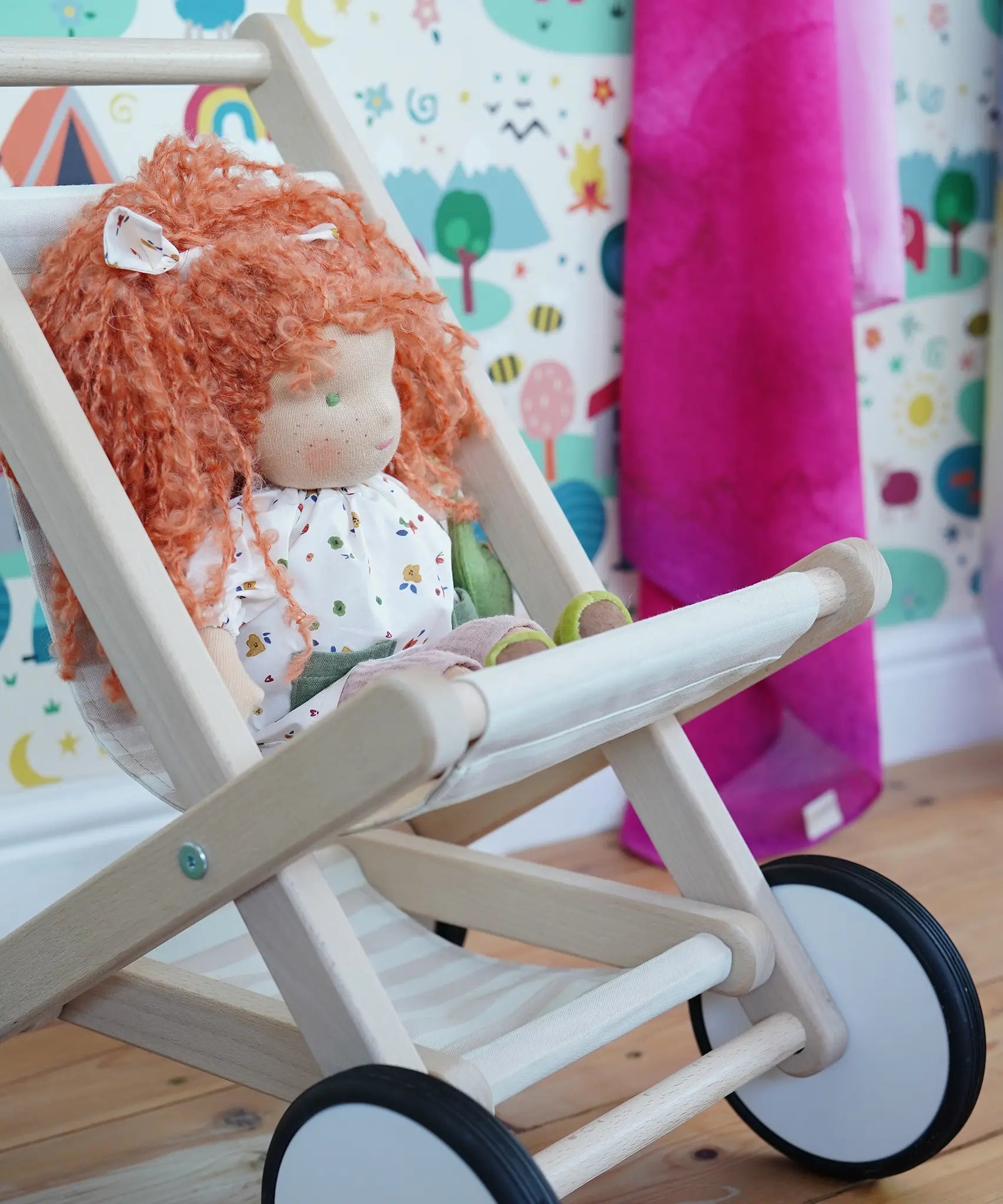 A wooden Bajo Toys stroller with a doll featuring curly orange hair and a white dress, set against a colorful children's room background with patterned wallpaper and a pink blanket.