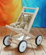 A  Bajo Toys wooden toy stroller with white wheels and a beige Senger teddy bear sitting inside, placed on a wooden floor against a colorful rainbow-striped backdrop.