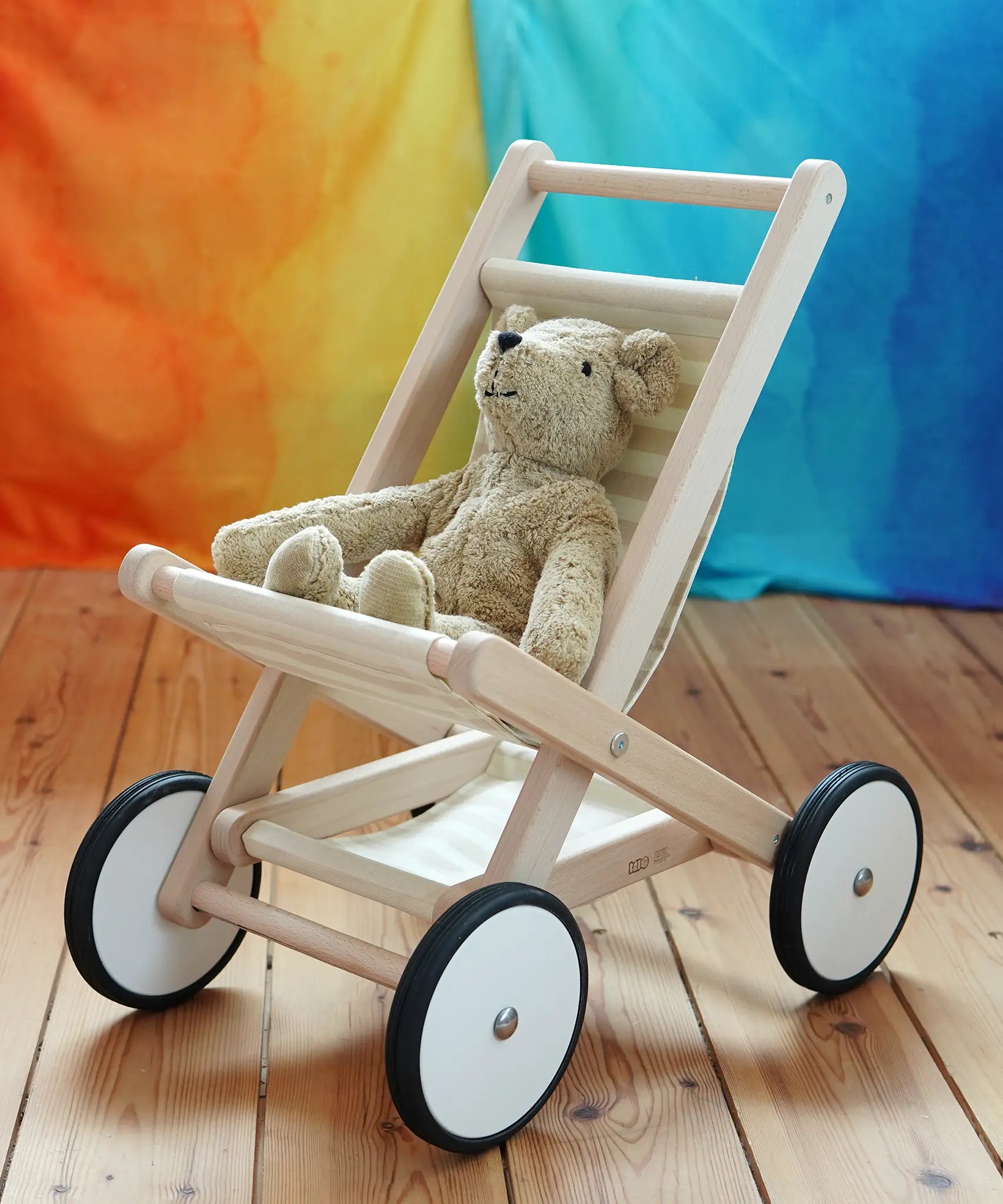A  Bajo Toys wooden toy stroller with white wheels and a beige Senger teddy bear sitting inside, placed on a wooden floor against a colorful rainbow-striped backdrop.
