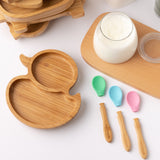 Eco Rascals Duck Bamboo Suction Plate, with a stack of other bamboo plates and bowl in the background, a glass of milk and detachable heads of the silicone and bamboo baby spoons. 