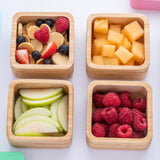 Four open bamboo snack pots with different fresh fruits inside with the colourful silicone lids next to them. 