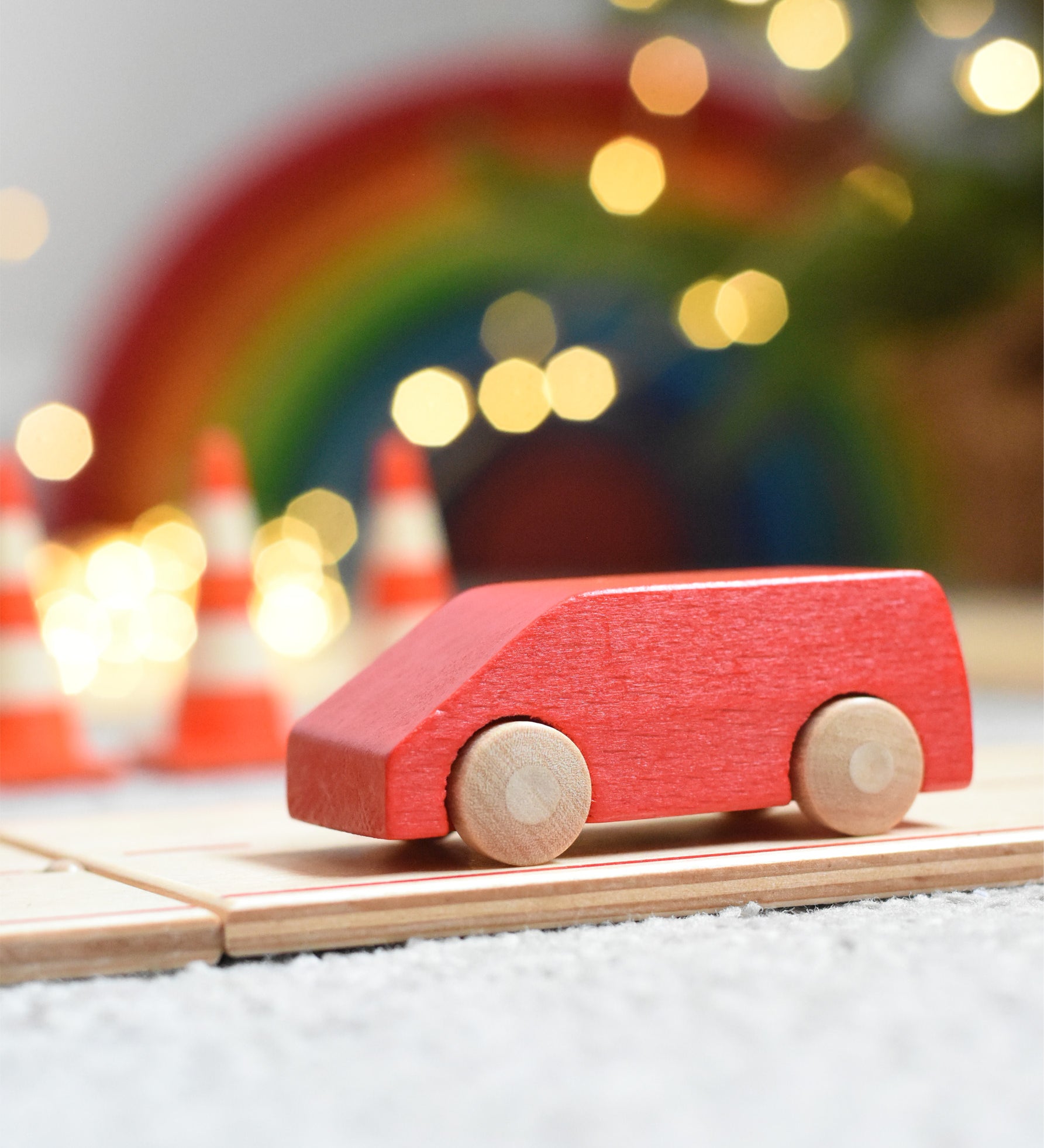 The Beck Holzspielzeug mini wooden toy van, driving along the wooden road track with traffic cones, twinkling lights, and a Grimm's rainbow in the background. 