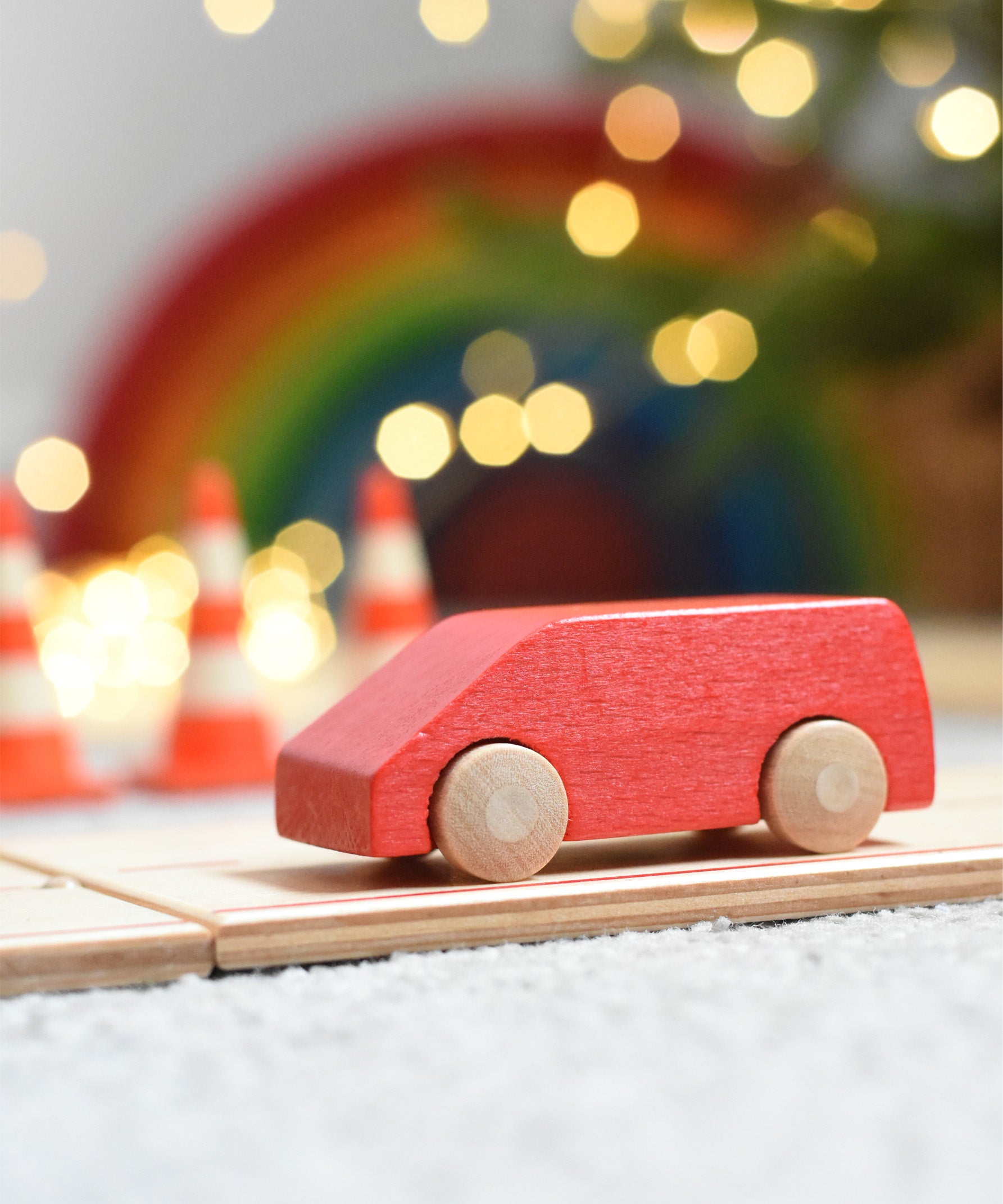 The Beck Holzspielzeug mini wooden toy van, driving along the wooden road track with traffic cones, twinkling lights, and a Grimm's rainbow in the background. 