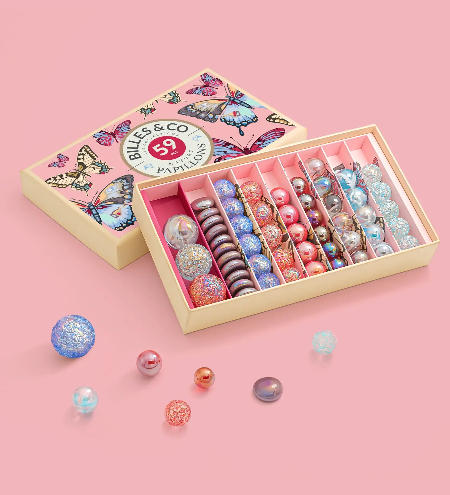 Different sized marbles from the Billes & Co butterfly anamorphic set on a pink surface with a pink butterfly box in the background.