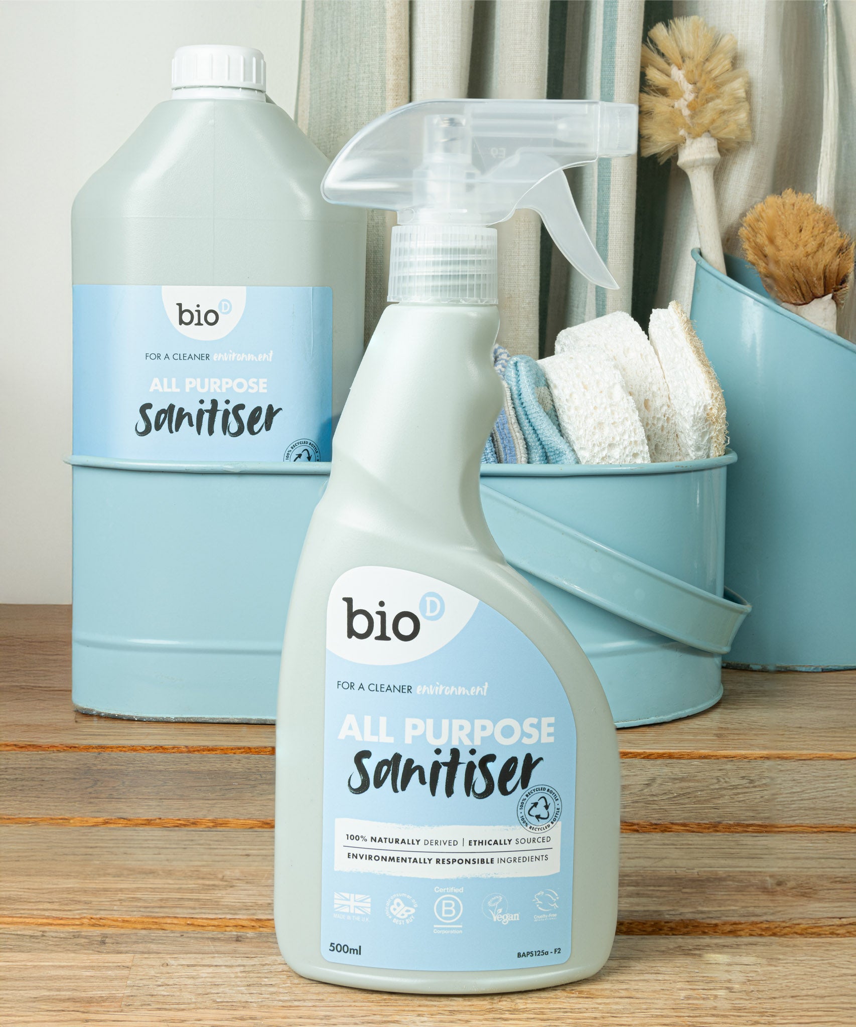 Bio-d 500ml all purpose sanitiser spray in fragrance free standing on a wooden surface with a 5L bottle in the background with cleaning products. This sanitiser spray is available at Babipur.