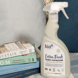A 500ml spray bottle of Bio-D vegan and cruelty free cotton fresh fabric refresher standing on a blanket next to books