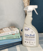 A 500ml spray bottle of Bio-D vegan and cruelty free cotton fresh fabric refresher standing on a blanket next to books
