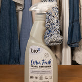 A 500ml spray bottle of Bio-D vegan and cruelty free cotton fresh fabric refresher standing on a wooden surface with clothes in the background