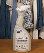 A 500ml spray bottle of Bio-D vegan and cruelty free cotton fresh fabric refresher standing on a wooden surface with clothes in the background
