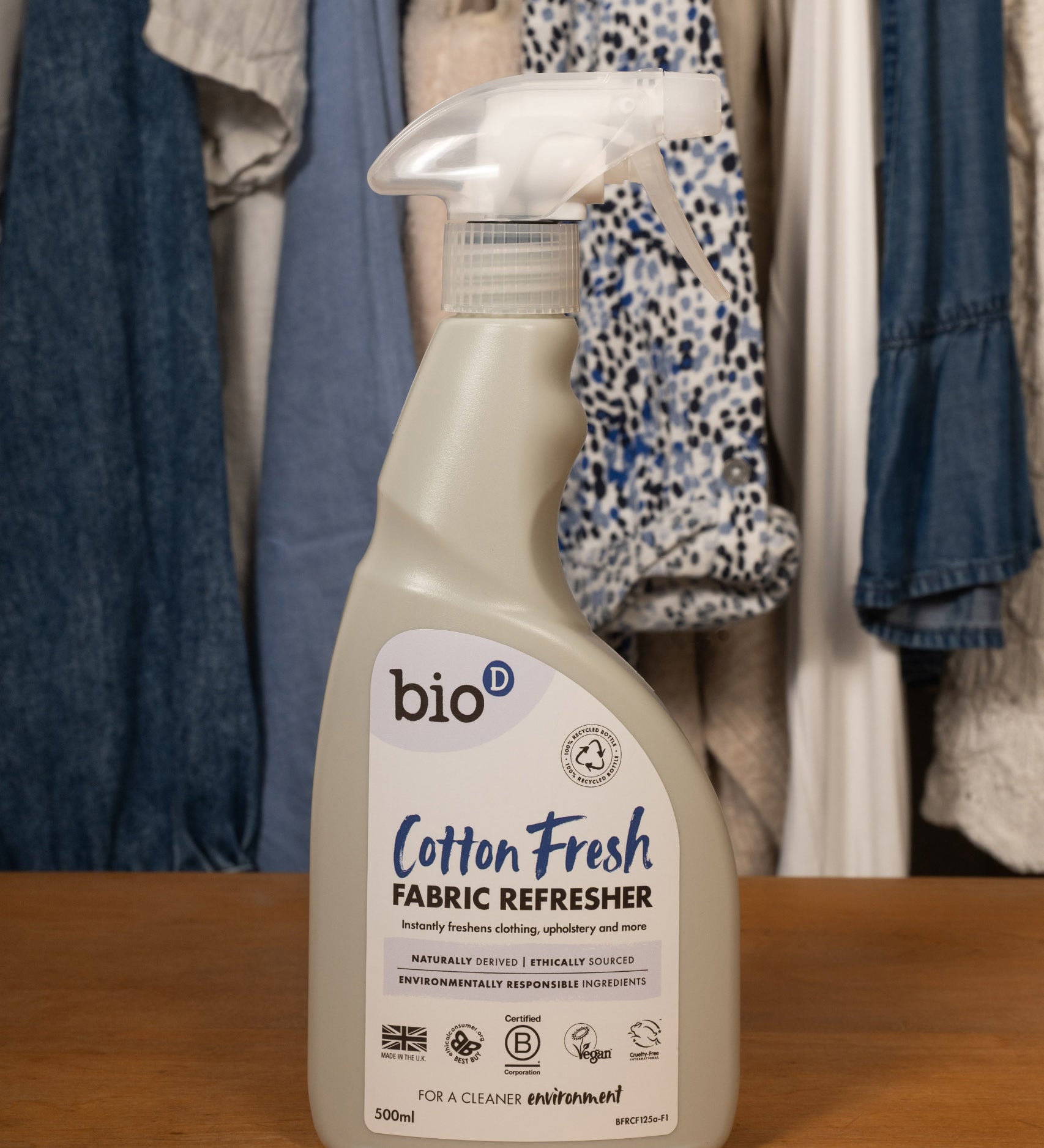 A 500ml spray bottle of Bio-D vegan and cruelty free cotton fresh fabric refresher standing on a wooden surface with clothes in the background