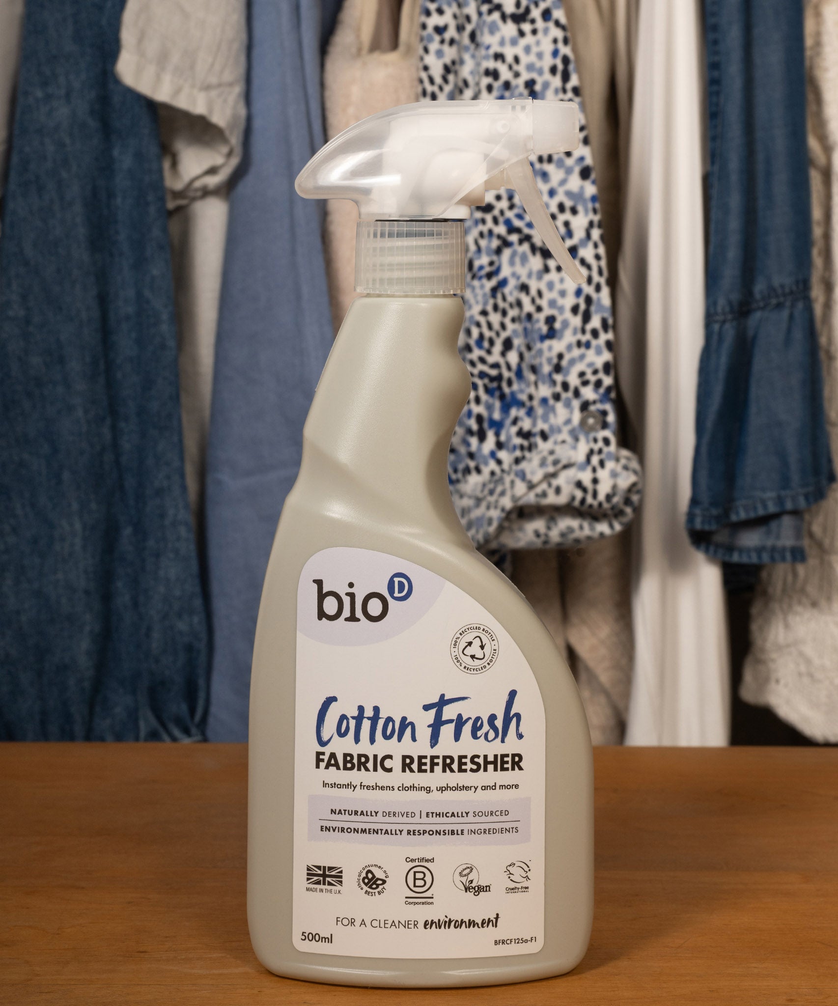 A 500ml spray bottle of Bio-D vegan and cruelty free cotton fresh fabric refresher standing on a wooden surface with clothes in the background
