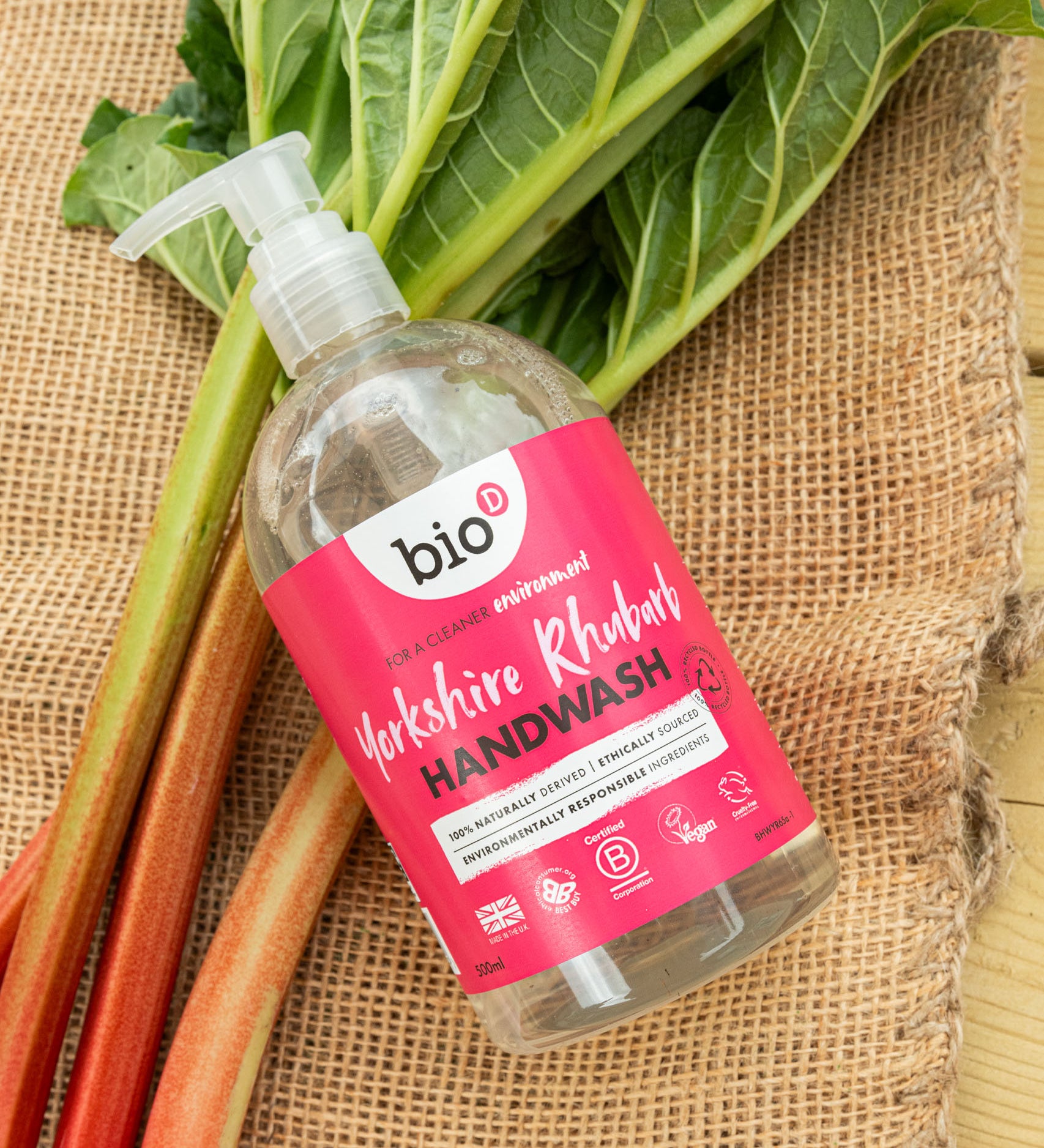 A flatlay image of a 500ml bottle of Bio-D cleansing hand wash  in Yorkshire rthubarb on top of rhubarb on a hessian sheet. This hand wash is available at Babipur.