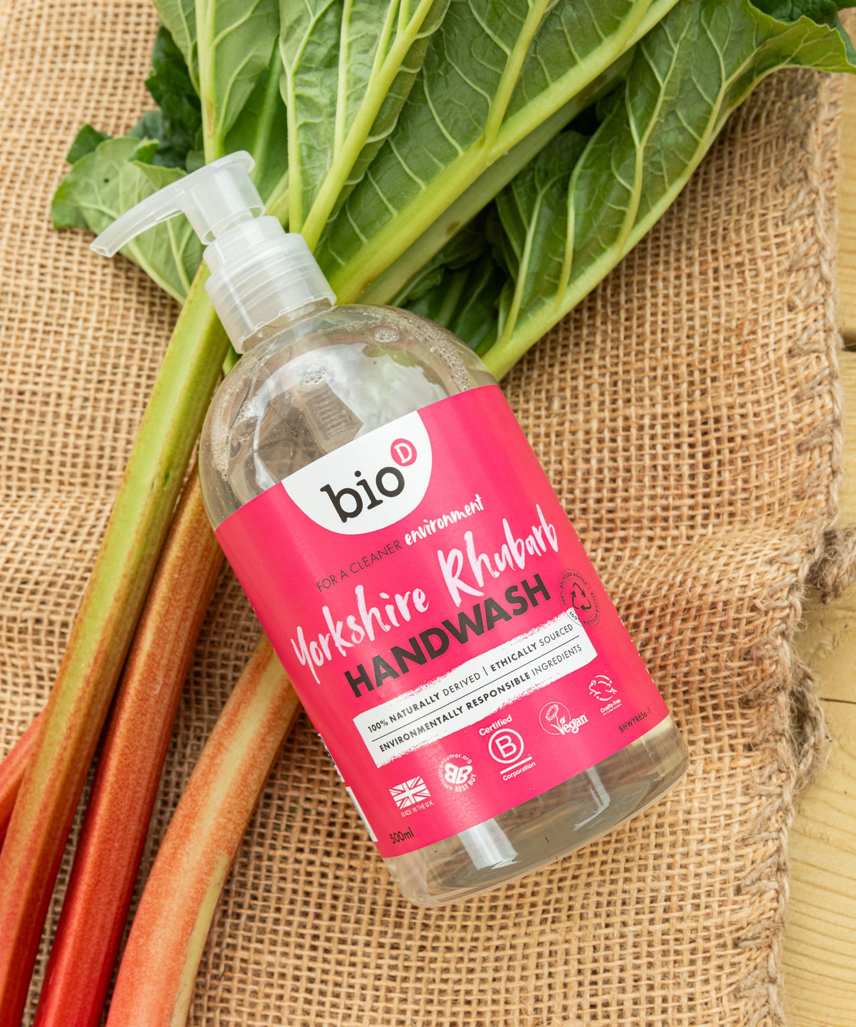 A flatlay image of a 500ml bottle of Bio-D cleansing hand wash  in Yorkshire rthubarb on top of rhubarb on a hessian sheet. This hand wash is available at Babipur.