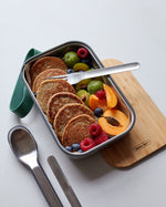 A close up of the Black + Blum stainless steel cutlery set next to a olive original Lunchbox with food inside