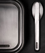 A close up of the Black + Blum stainless steel cutlery set next to a stainless steel lunchbox