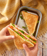A close up of a adult hand holding a toasted sandwich with another in a Black + Blum stainless steel airtight stainless steel sandwich box 