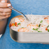 A close up and adult holding the fork from the Black + Blum stainless steel cutlery set and eating from a stainless steel lunchbox