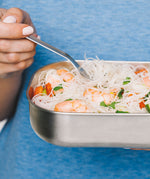 A close up and adult holding the fork from the Black + Blum stainless steel cutlery set and eating from a stainless steel lunchbox