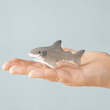 An adult hand holding the Bumbu handmade and hand painted wooden grey baby shark figure showing size