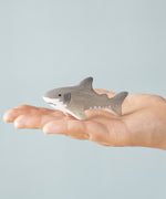 An adult hand holding the Bumbu handmade and hand painted wooden grey baby shark figure showing size